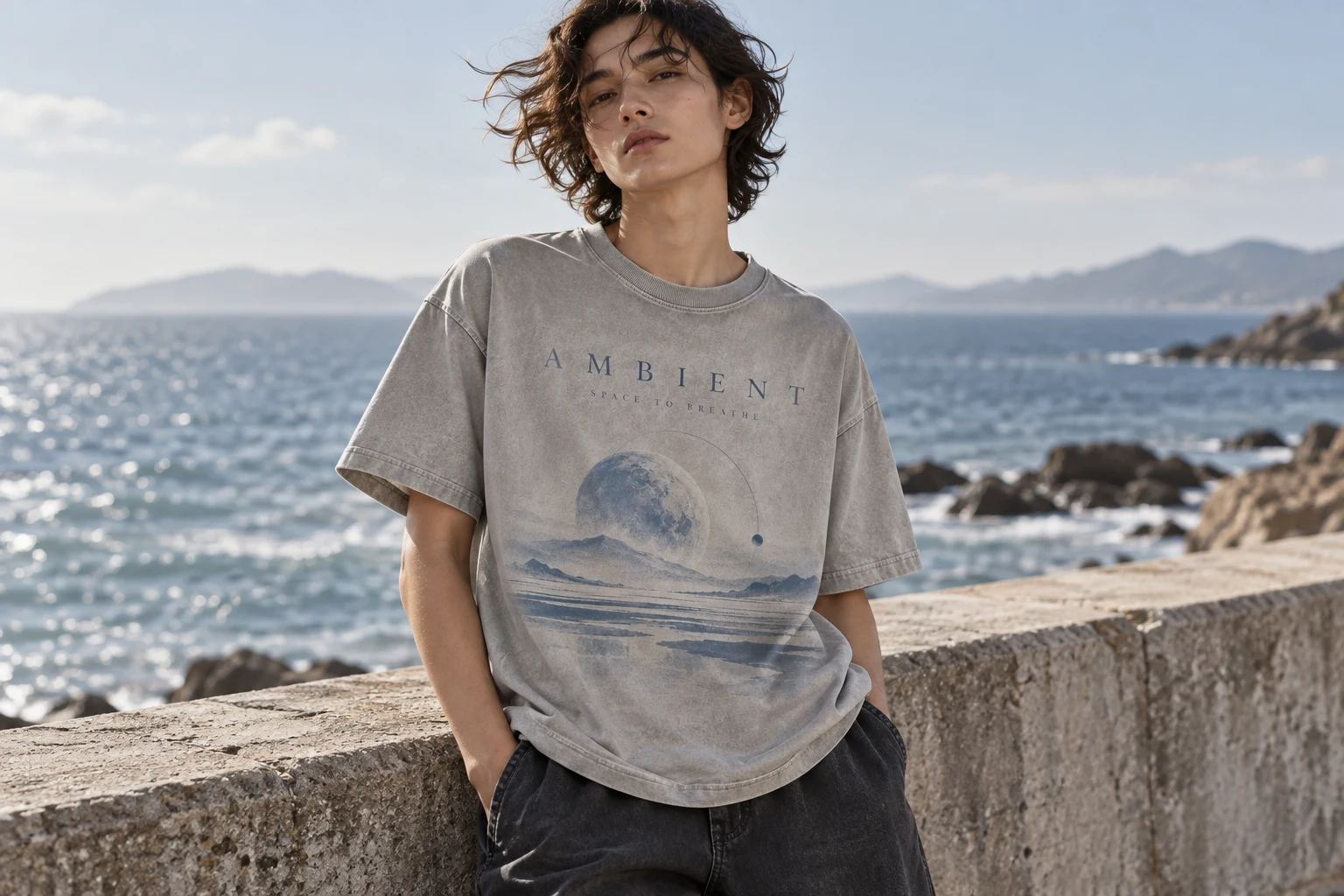 Adult model in the stone-grey Ambient T-shirt beside a calm seaside promenade